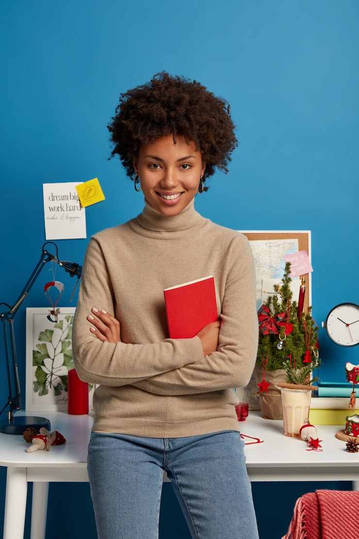 Photo gratuit tir vertical de l'enseignante aux cheveux bouclés heureux se prépare pour les leçons à la maison, tient le manuel rouge, pose contre le bureau