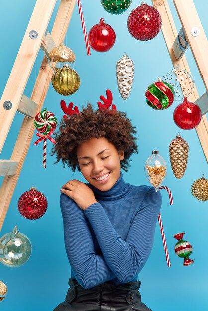 Tir vertical de la femme afro-américaine heureuse bénéficie d'une atmosphère magique festive à la maison porte des poses confortables à col roulé