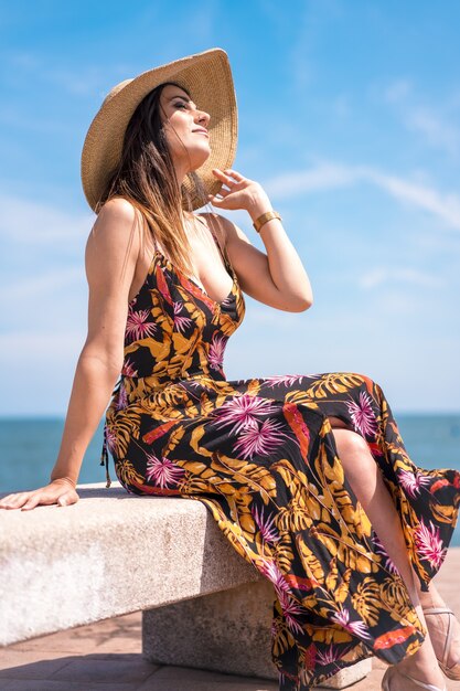 Tir vertical d'une femme dans une robe d'été fleurie et un chapeau assis au bord de la mer capturé en Espagne