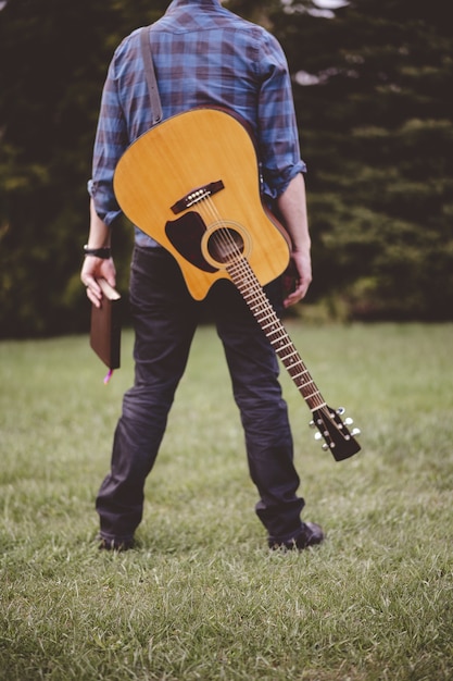 Photo gratuite tir vertical d'un homme avec une guitare et un livre dans une main debout sur l'herbe