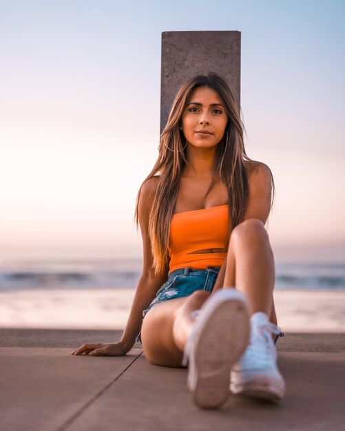 Tir vertical d'une jeune femme brune en short jeans et un haut orange assis sur la plage