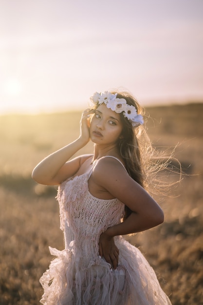 Tir vertical d'une jeune femme de race blanche en robe blanche et couronne de fleurs blanches posant dans un champ