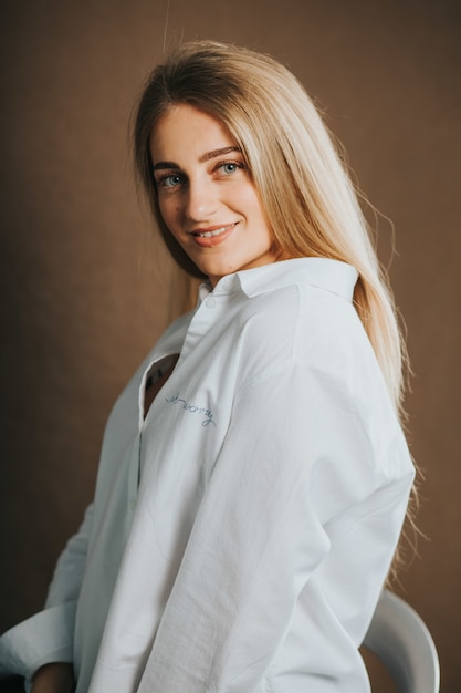 Tir vertical d'une jolie femme blonde dans une chemise blanche posant sur un mur marron