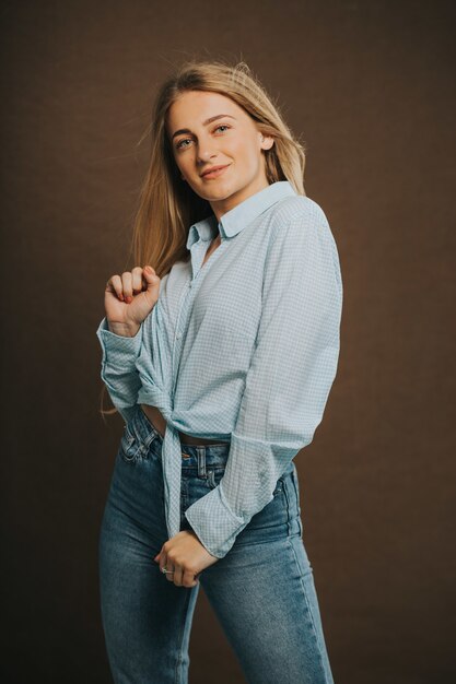 Tir vertical d'une jolie femme blonde en jeans et une chemise courte posant sur un mur marron