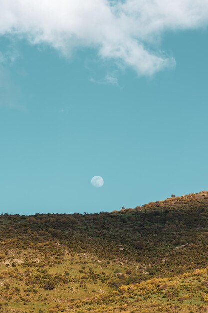 Tir vertical d'une pleine lune sur un ciel bleu au-dessus d'une colline pendant la journée