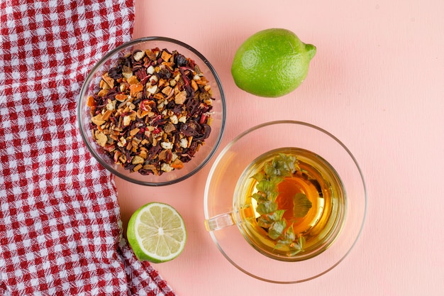 Tisane dans une tasse en verre avec limes, herbes séchées à plat sur rose et torchon