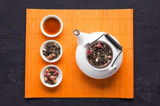 Tisane et son ingrédient disposés en rang avec la théière sur un napperon orange sur un fond noir