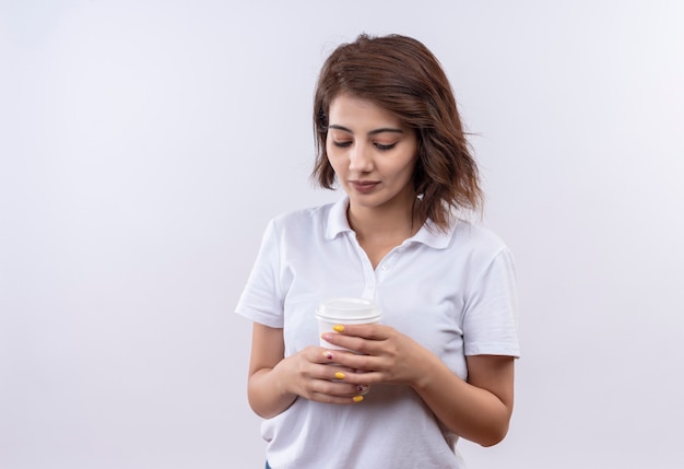Titmid jeune fille aux cheveux courts portant un polo blanc tenant une tasse de café à la bas