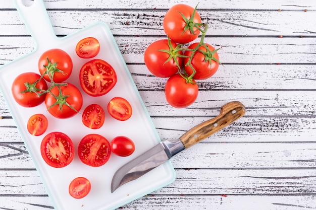 Photo gratuite tomate pleine et demi sur une planche à découper blanche des branches de tomate près du couteau à découper sur une surface en bois blanche