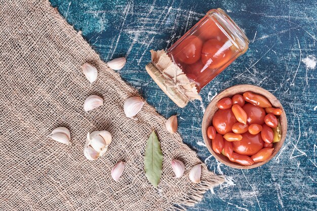 Tomates marinées dans un bocal en verre et bol avec de l'ail sur bleu.