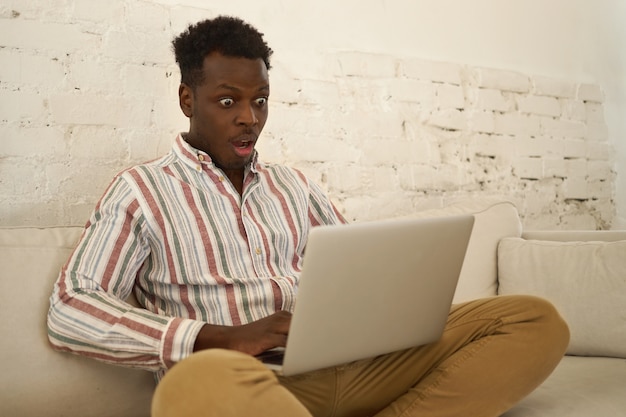 Étonné drôle jeune homme afro-américain assis sur un canapé avec les jambes pliées keepig bouche ouverte tout en lisant des nouvelles inattendues