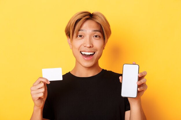 Étonné heureux mec asiatique montrant la carte de crédit et l'écran du smartphone, souriant fasciné, debout mur jaune.