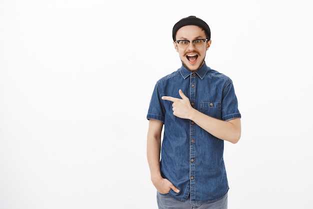 Étonné et impressionné beau mec adulte barbu en chemise bleue et bonnet noir pointant vers la gauche avec l'index et souriant largement d'excitation et de joie d'être ravi