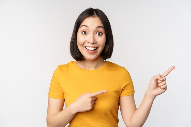 Étonné jeune femme asiatique montrant la publicité de côté pointant du doigt le logo de la marque de texte de promotion debout heureux sur fond blanc