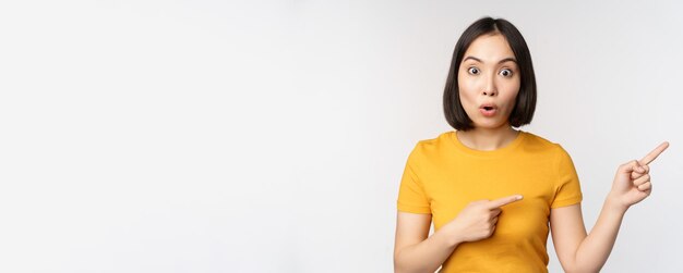 Étonné jeune femme asiatique montrant la publicité de côté pointant du doigt le logo de la marque de texte de promotion debout heureux sur fond blanc