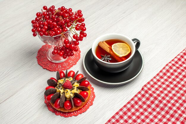 Top close view groseille rouge dans un verre de cristal sur le napperon en dentelle ovale rouge et une tasse de thé citron cannelle et nappe à carreaux rouge-blanc sur la table en bois blanc