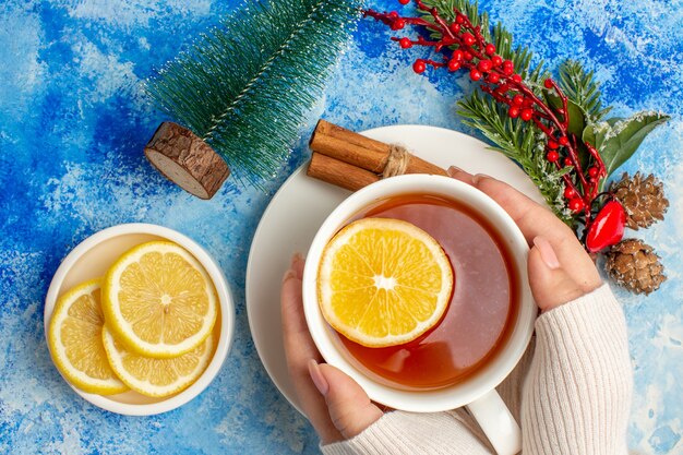 Top vue rapprochée tasse de thé dans les mains des femmes branche d'arbre de Noël coupé des citrons sur une soucoupe sur une table bleue