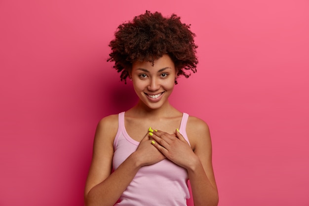 Touché et ravi, la belle femme entend une histoire réconfortante, serre les mains contre le cœur, se sent reconnaissant, vous promet, sourit doucement, porte des vêtements décontractés, apprécie les beaux cadeaux, pose sur un mur rose.