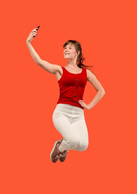 Toujours sur mobile. Sur toute la longueur de la jolie jeune femme prenant le téléphone et faisant selfie en sautant sur fond de studio rouge. Mobile, mouvement, mouvement, concepts commerciaux