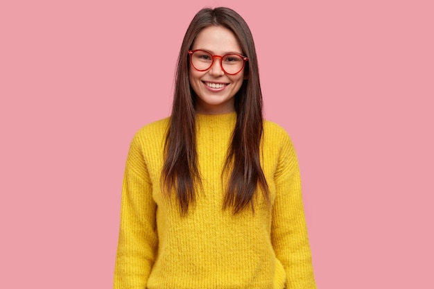 Tour De Taille D'une Jolie Jeune Femme Brune Satisfaite Avec Une Expression Heureuse, Porte Des Lunettes Et Un Pull Jaune, Des Modèles Sur Fond Rose