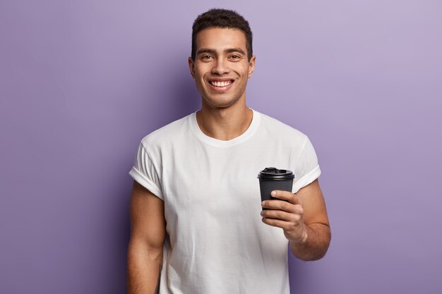 Tour de taille de sourire joyeux beau mec tient du café à emporter en papier, aime le temps libre avec un ami, boit des boissons aromatiques, porte un t-shirt blanc maquette, des modèles contre le mur violet