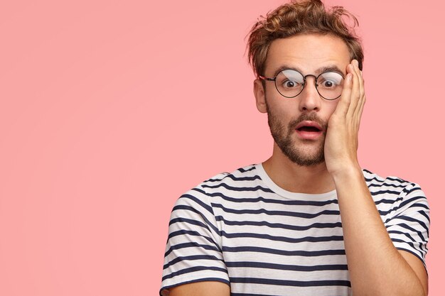 Étourdi, un regard masculin bouclé et agréable avec étonnement, garde la main sur la joue, a une réaction inattendue, vêtu d'un t-shirt rayé