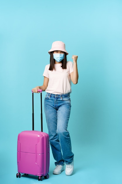 Touriste asiatique enthousiaste de fille dans le masque médical tenant la valise pleine longueur voyageant à l'étranger ...