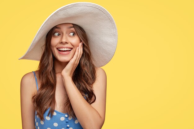 touriste assez heureux, se réjouit d'une journée pleine de miracles et de moments inoubliables, porte des vêtements d'été, regarde joyeusement de côté, garde la main sur la joue, modèles sur un mur jaune avec espace de copie