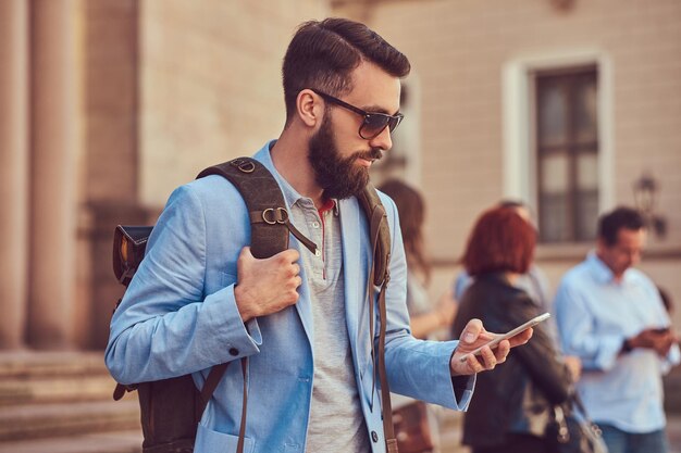 Un touriste avec une barbe complète et une coupe de cheveux, portant des vêtements décontractés et des lunettes de soleil, tient un sac à dos et envoie des SMS sur un smartphone, debout dans une rue antique, pendant l'excursion en Europe.