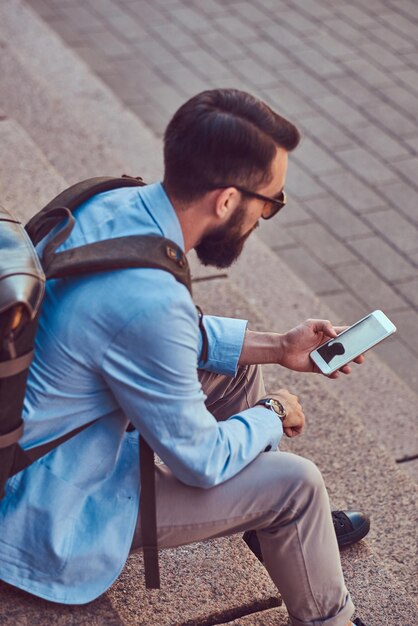 Un touriste avec une barbe pleine et une coupe de cheveux, portant des vêtements décontractés avec un sac à dos, envoyant des SMS sur un smartphone, assis sur une marche dans une rue antique.