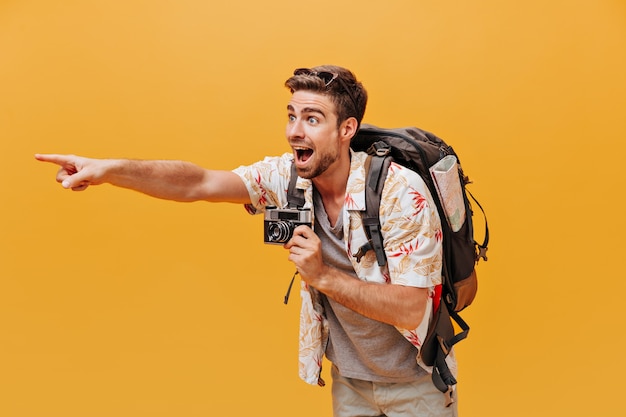 Touriste brillant avec barbe de gingembre en chemise blanche et t-shirt gris montrant de côté son doigt et tenant la caméra sur un mur isolé