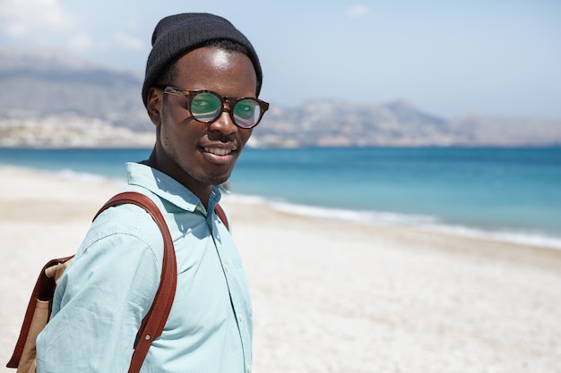 Touriste homme noir à la mode attrayant vêtu de vêtements et d'accessoires à la mode posant contre l'eau bleue et le sable blanc
