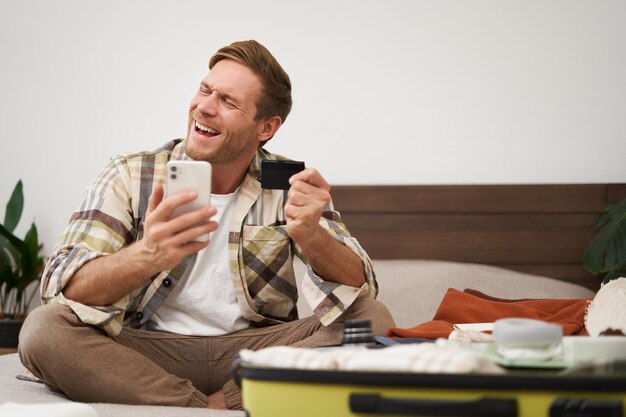 Photo gratuite un touriste joyeux avec une carte de crédit et un téléphone portable payant des billets de vacances assis sur le lit