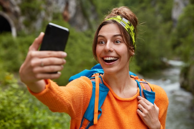 Touriste joyeux avec un large sourire, tient un téléphone portable devant, fait un portrait de selfie