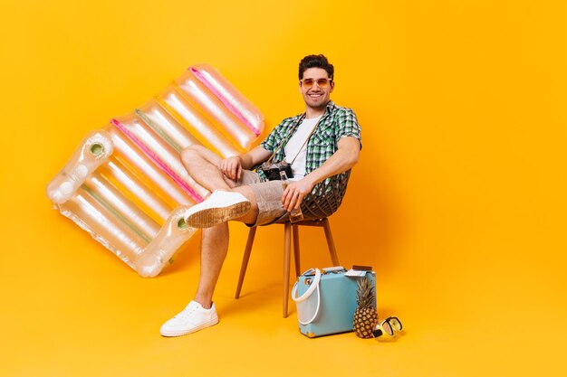 Touriste masculin dans des verres orange se détend assis sur une chaise et buvant de la bière Un gars en chemise verte pose avec un matelas gonflable de valise et un ananas sur fond orange