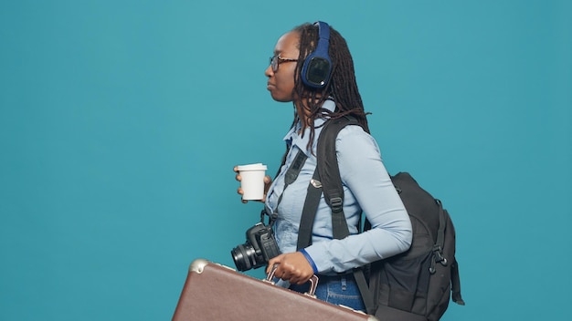 Touriste partant en vacances avec valise et sac à dos, partant en vacances avec appareil photo pour prendre des photos. Photographe écoutant de la musique sur des écouteurs et buvant une tasse de café.