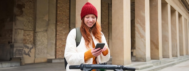 Photo gratuite une touriste rousse avec sac à dos utilise un téléphone portable pour louer un scooter dans les rues de la ville européenne