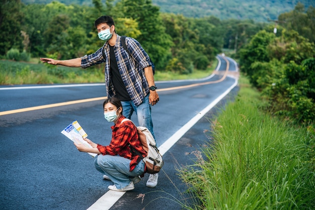 Les touristes féminines s'assoient et regardent la carte, les touristes masculins faisant semblant de faire de l'auto-stop. Les deux portent des masques et sont sur le bord de la route.