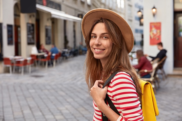 Tourné à l'extérieur d'un touriste européen joyeux se promène dans les rues de la ville, porte un sac à dos, porte un chapeau marron, un pull rayé