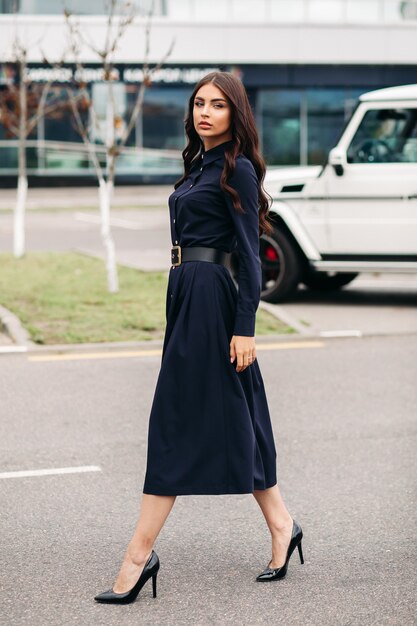 Tourné sur toute la longueur de la belle jeune femme élégante vêtue d'une robe noire et marchant dans la rue de la ville. Concept de style et de mode