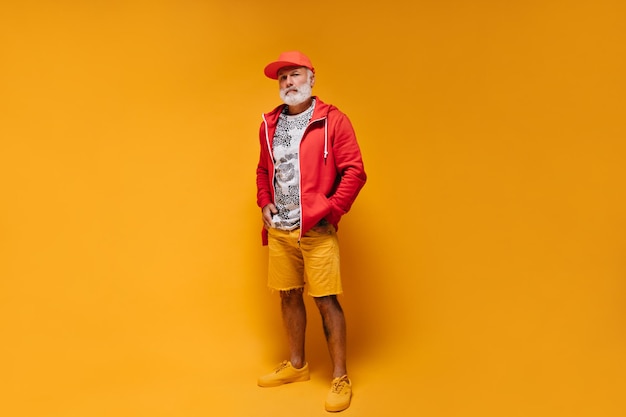 Tourné sur toute la longueur de l'homme en short orange et veste rouge gars élégant avec barbe en t-shirt imprimé léopard blanc à capuche élégant et casquette posant