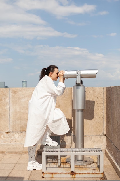 Photo gratuite toursit femme caucasienne debout sur la tour du bâtiment regardant à travers un télescope de jumelles bénéficiant d'une vue panoramique sur la ville métropolitaine, paysage avec des bâtiments urbains depuis le point d'observation,