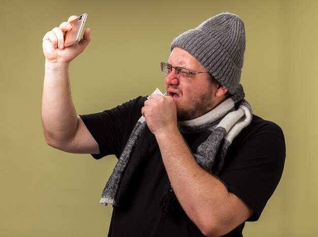 Toussant un homme malade d'âge moyen portant un chapeau d'hiver et une écharpe prend un selfie isolé sur un mur vert olive