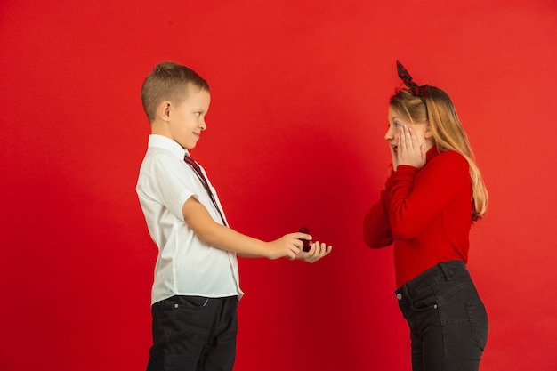 Tout comme un adulte. Célébration de la Saint-Valentin, enfants caucasiens heureux et mignons isolés sur fond de studio rouge. Concept d'émotions humaines, expression faciale, amour, relations, vacances romantiques.