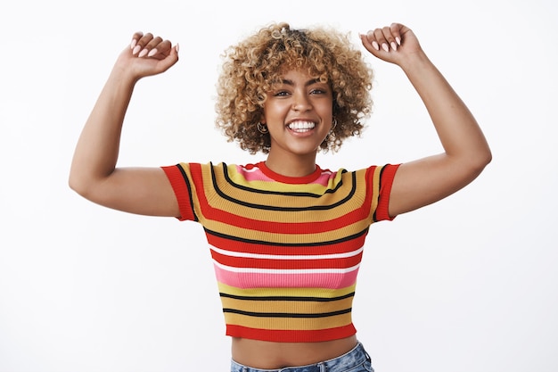 Tout le monde lève les mains en l'air. Portrait d'une jeune fille afro-américaine joyeuse et insouciante s'amusant à lever les bras haut et à danser souriant largement, se sentant énergique et optimiste sur un mur blanc