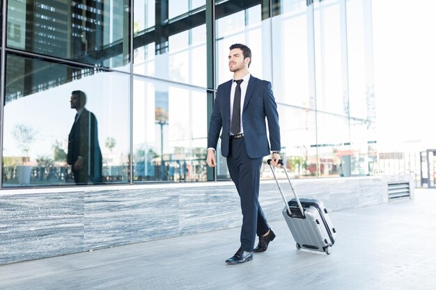 Toute la longueur du beau jeune homme en costume marchant avec des bagages à l'extérieur de l'immeuble de bureaux