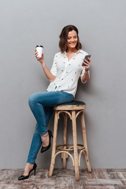 Toute la longueur de femme souriante posant sur une chaise avec une tasse de café à la main et à l'aide de smartphone sur gris