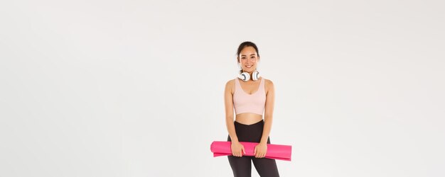 Toute la longueur de la fille asiatique mince souriante avec tapis en caoutchouc pour les exercices de fitness casque regardant c