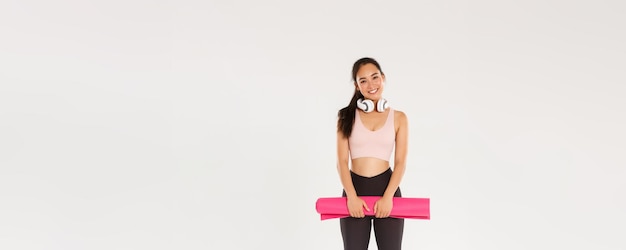 Toute la longueur d'une jolie fille asiatique comme la forme physique tenant un tapis en caoutchouc pour des exercices ou des cours de yoga debout