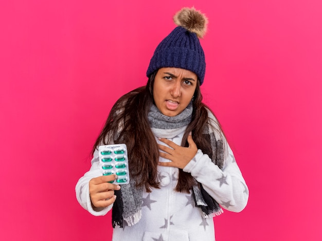 Toux jeune fille malade portant chapeau d'hiver avec écharpe tenant des pilules mettant la main sur le coeur isolé sur rose